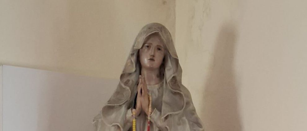 Eine Statue einer Frau mit einem Schleier steht auf einem Regal. Die Statue hat einen Rosenkranz und ein Kreuz. Vor der Statue stehen zwei rote Kerzen.