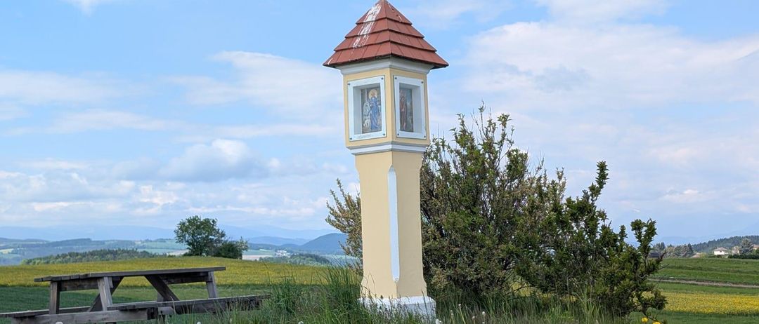 Ein hoher Betonpfeiler mit rotem Dach und kleinen Fenstern steht in einem Grasfeld. Ein hölzerner Picknicktisch ist in der Nähe.