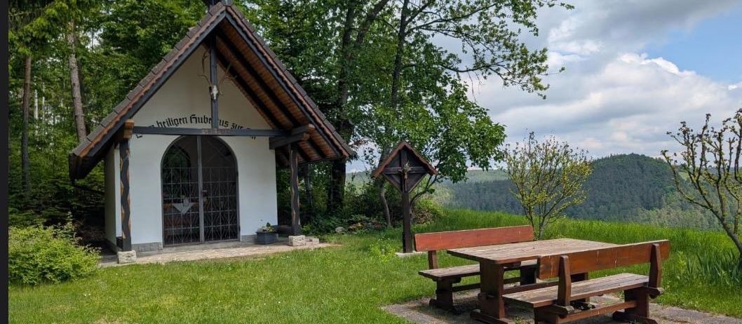 Eine kleine Kapelle mit einem Kreuz oben, inmitten grüner Umgebung. Eine Holzbank und ein Tisch stehen in der Nähe.