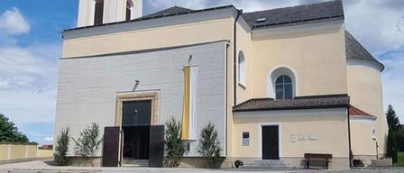 Eine beigefarbene Kirche mit einem roten Dach und einem spitzen Turm steht unter einem bewölkten Himmel. Eine gelbe Fahne hängt an der Wand.