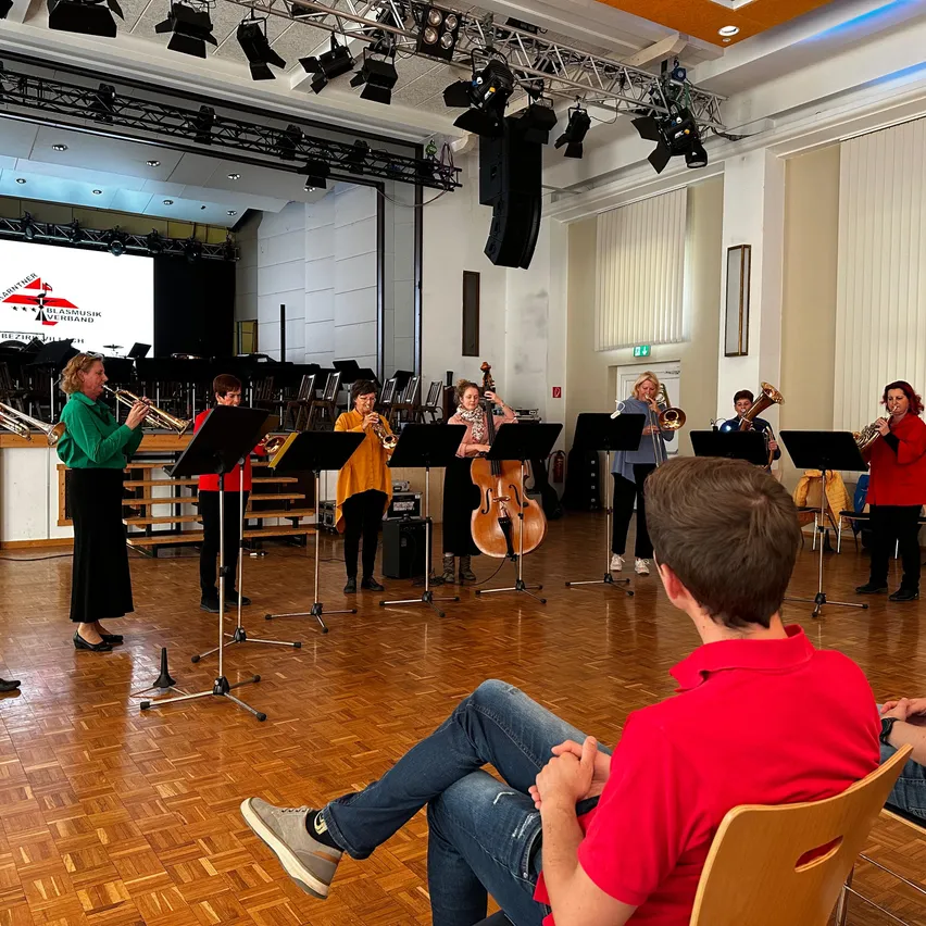 Eine Gruppe von Musikern spielt Trompeten, Celli und andere Instrumente in einem Raum mit einer großen Leinwand im Hintergrund, während ein Mann auf einem Stuhl im Vordergrund sitzt.