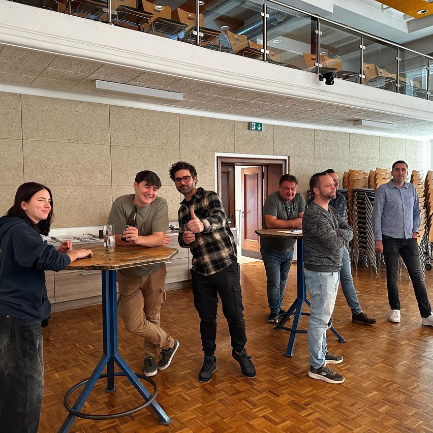 Eine Gruppe von Menschen in Freizeitkleidung versammelt sich um Tische in einem geräumigen Raum mit Holzböden. Ein Mann gibt Daumen hoch, und andere lächeln. Dahinter öffnet sich eine Tür, und ein Balkon mit Stühlen ist sichtbar.