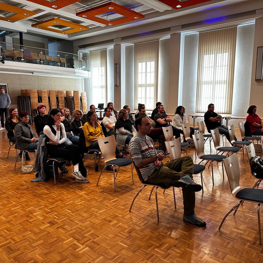 Eine Gruppe von Menschen sitzt in Stühlen in einem großen Raum, einige schauen zu einem Mann, der sich in der Nähe einer Balkonbrüstung befindet.