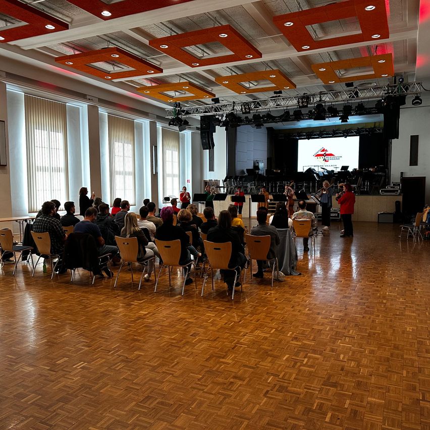 Eine Gruppe von Menschen sitzt in Stühlen in einem Raum mit Bühne, Musikern und einer großen Leinwand mit Logo.