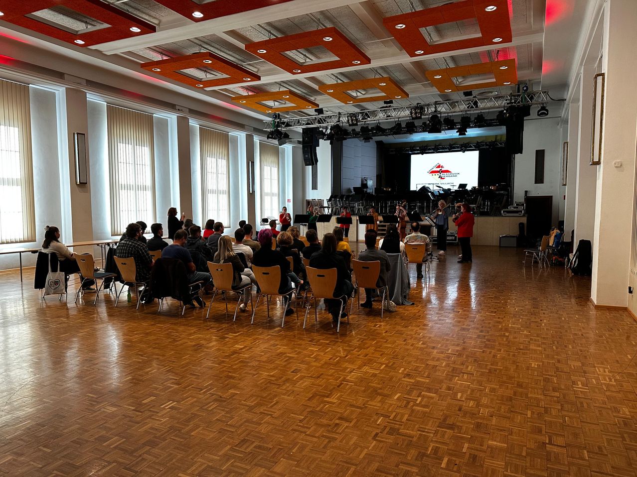 Eine Gruppe von Menschen sitzt in Stühlen in einem Raum mit Bühne, Musikern und einer großen Leinwand mit Logo.