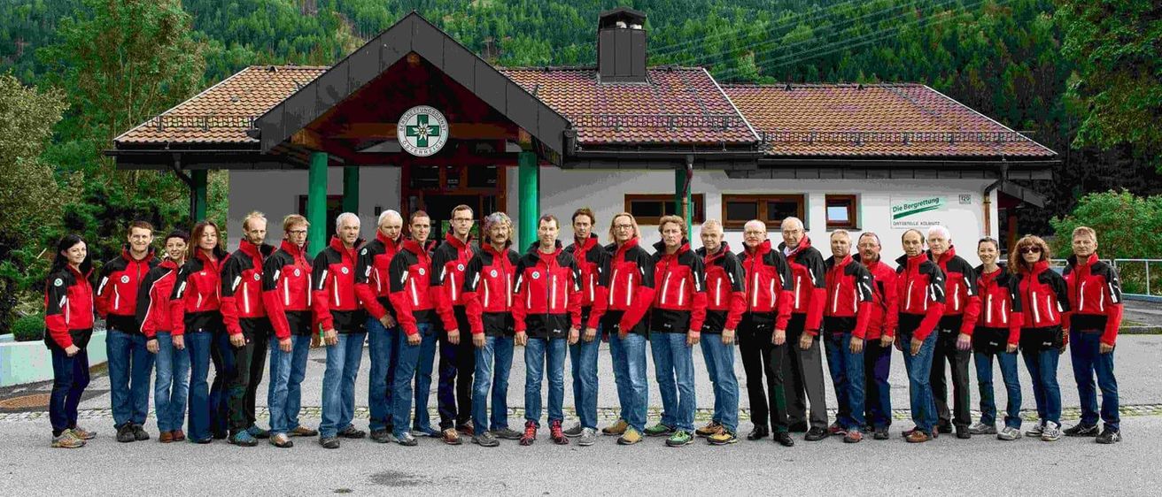 Eine Gruppe von Menschen in roten und schwarzen Jacken steht vor einem Gebäude mit einem Berg im Hintergrund.