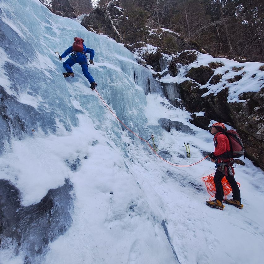 Zwei Personen klettern einen vereisten Berg hinauf, eine in Blau und eine in Rot, beide mit Rucksäcken und Sicherheitsseilen ausgestattet. Schnee bedeckt den Boden und die Felsen.