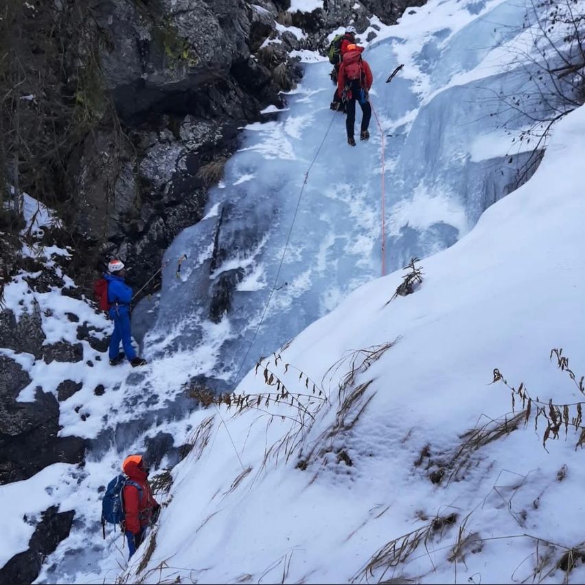 Drei Personen klettern einen vereisten Berg hinauf, zwei in roten Jacken und Rucksäcken, eine in einer blauen Jacke. Sie benutzen Seile zur Sicherheit, umgeben von Schnee und Felsen.