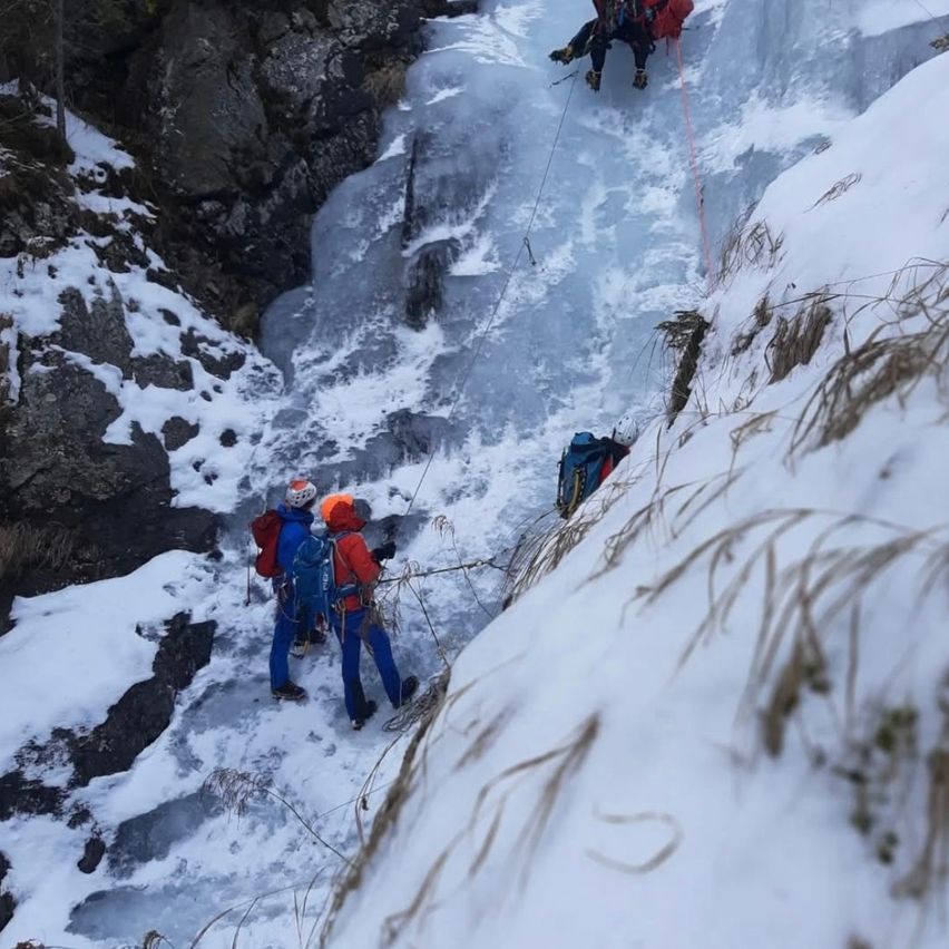 Eine Gruppe von Kletterern erklimmt einen schneebedeckten Fels, gesichert mit Seilen. Zwei Kletterer sind im Vordergrund, während ein weiterer höher positioniert ist. Sie sind alle mit Helmen und Rucksäcken ausgestattet.