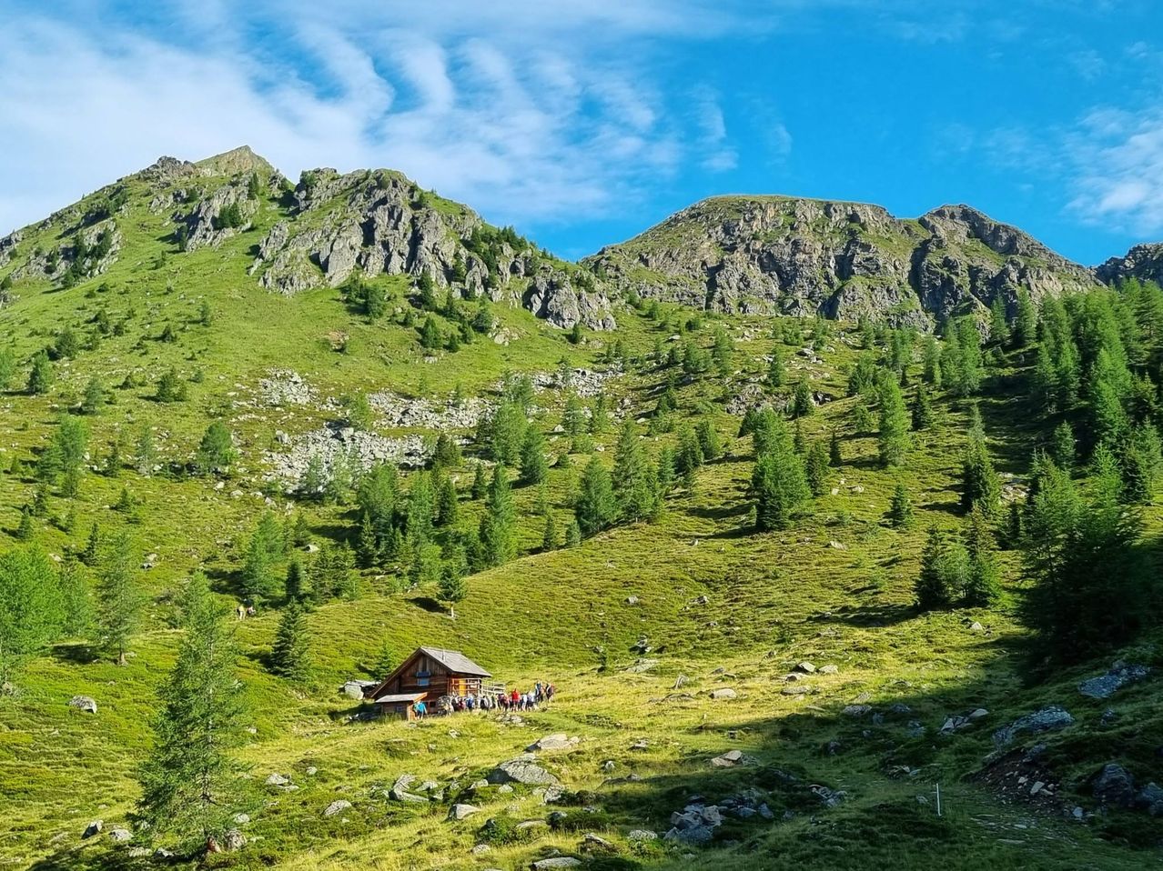 Eine kleine Holzhütte steht auf einem grasigen Hang mit Kiefern und Felsvorsprüngen, unter einem blauen Himmel mit verstreuten Wolken. Eine Gruppe von Menschen ist vor der Hütte versammelt.