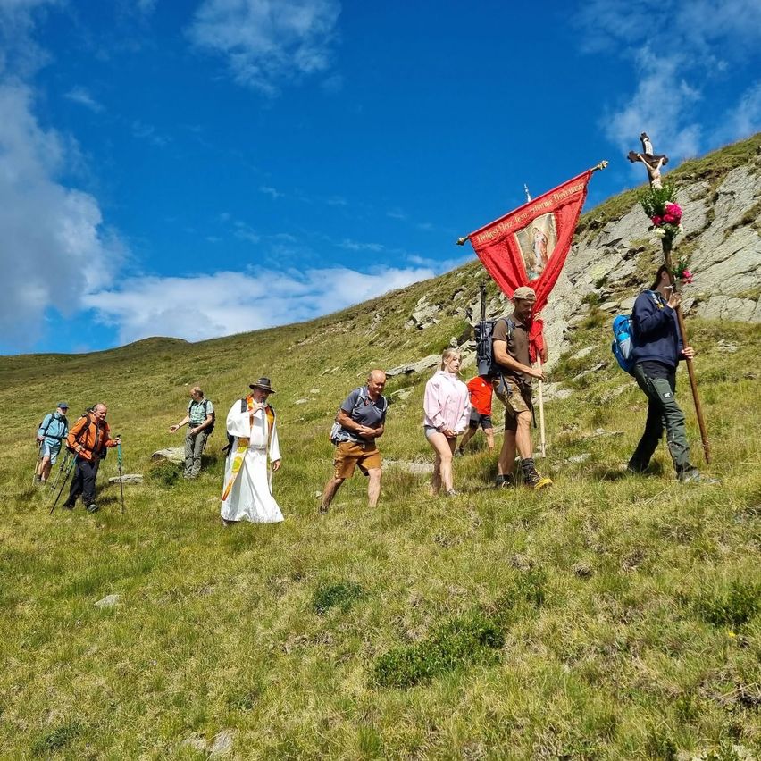 Eine Gruppe von Wanderern, einschließlich eines Priesters in Weiß, wandert auf einem grasbewachsenen Hügel mit einem roten Banner und einem Kreuz an einem Stab.