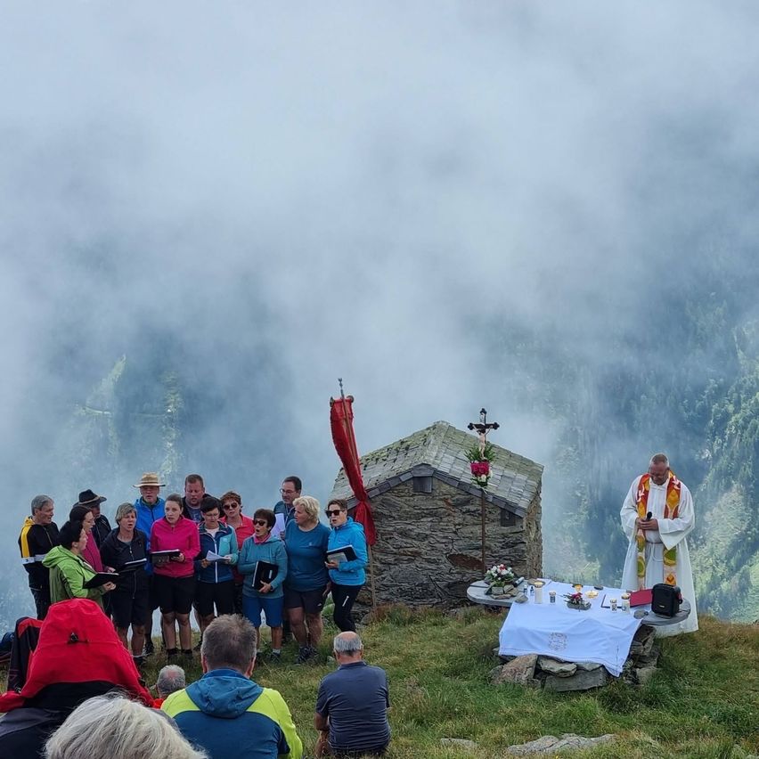 Eine Gruppe von Menschen in Outdoor-Kleidung steht in einem Kreis um einen Priester, der ein Mikrofon und ein Buch in der Hand hält. Sie befinden sich auf einem Grashügel mit einer kleinen Steinkapelle im Hintergrund.