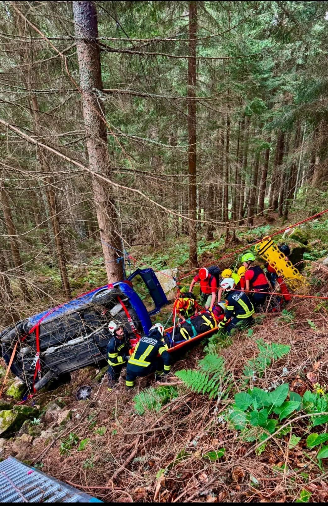 Rettungskräfte in roten und gelben Uniformen retten eine Person aus einem umgekippten Auto im Wald.
