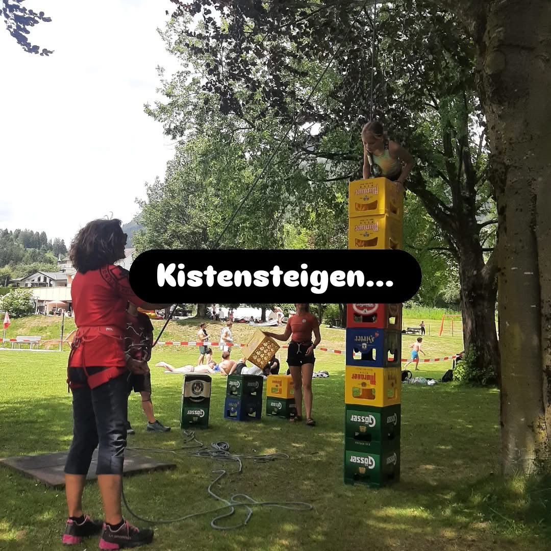 Menschen in roten Shirts klettern auf gestapelte Kisten in einem Park. Eine Person ist auf dem Stapel und eine andere klettert hoch. Die Kisten sind gelb und grün. Das Wort 'Kistensteigen' steht in der Mitte.
