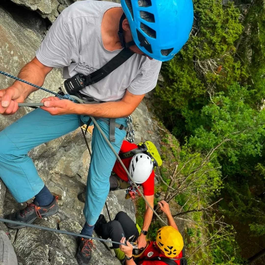 Ein Mann in Kletterausrüstung klettert eine Felswand hinauf, während eine andere Person ihm von unten hilft.
