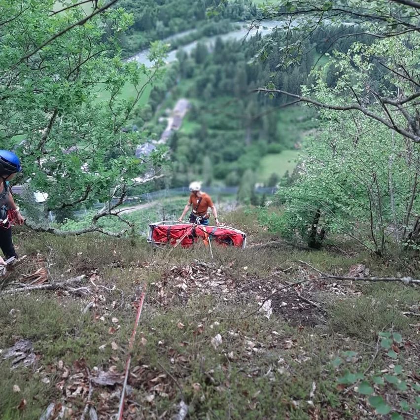Zwei Personen mit Helmen, eine mit einer roten Trage, bewegen sich einen bewaldeten Hang hinauf. Die Landschaft besteht aus dichten Bäumen, einem Fluss und entfernten Gebäuden.