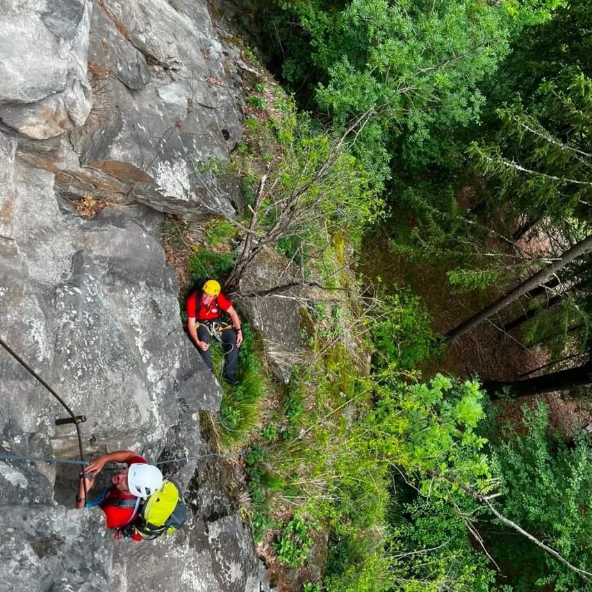 Zwei Kletterer in Sicherheitsausrüstung klettern einen steilen Felsvorsprung hinauf. Einer sitzt auf einer Kante, der andere klettert. Sie sind von dichter Waldvegetation umgeben.
