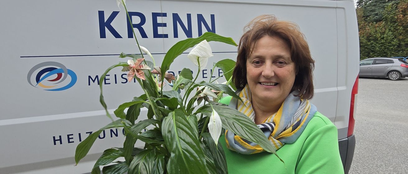 Eine Frau in einem grünen Shirt steht vor einem Krenn-Van und hält eine Topfpflanze mit weißen Blüten.