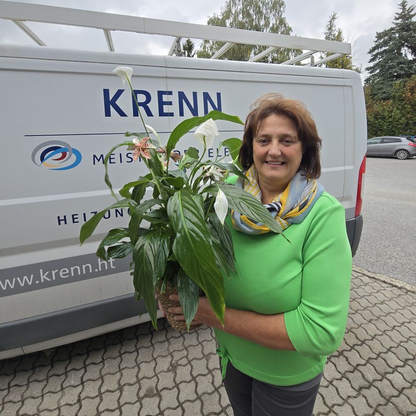 Eine Frau in einem grünen Oberteil hält eine Topfpflanze mit weißen Blüten vor einem Krenn-Servicewagen.