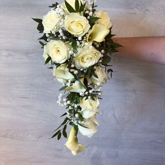 Ein Strauß weißer Rosen mit grünen Blättern und Babys Atemblumen, gehalten von einer Hand vor einem grauen Hintergrund.