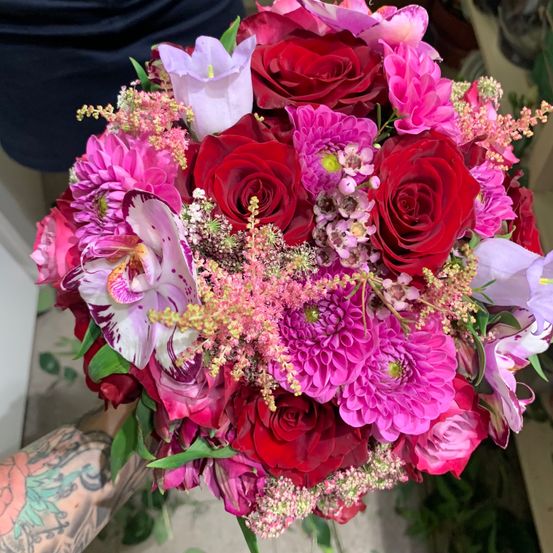 Ein leuchtender Blumenstrauß mit roten Rosen, pinken Dahlien und lila Orchideen, gehalten von einer Person mit Tätowierungen am Arm.