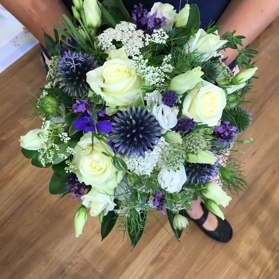 Eine Person hält einen Strauß aus weißen Rosen, blauen Disteln und grünen Blättern auf einem Holzboden.