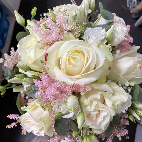 Ein Strauß weißer Rosen mit rosa Akzenten und verschiedenen Grünpflanzen, einschließlich Eukalyptusblättern, ist zu sehen. Die Rosen sind eng angeordnet, und das gesamte Farbschema ist sanft und elegant.