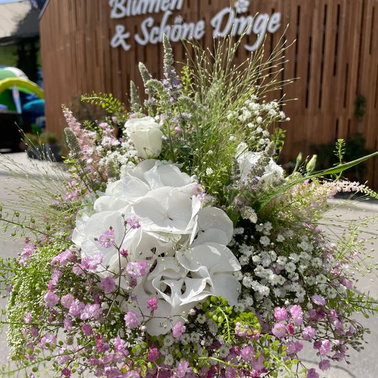 Ein großer Blumenstrauß aus weißen und rosa Blumen mit Grün vor einem Gebäude namens Blumen & Schöne Dinge.