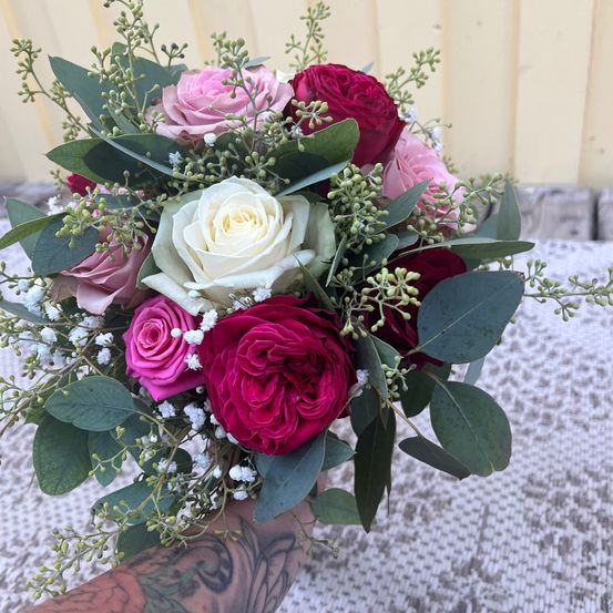 Ein Nahaufnahme eines Blumenarrangements mit rosa, weißen und roten Rosen, ergänzt durch grüne Blätter und kleine weiße Blüten, gehalten von einer tätowierten Hand.