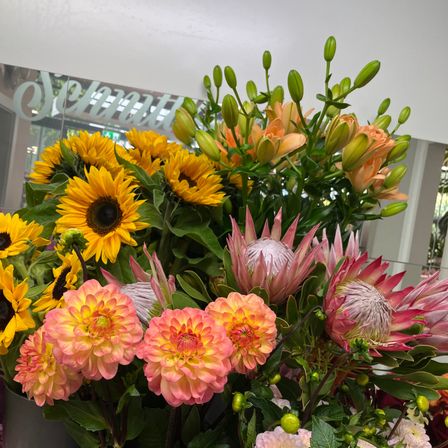 Ein lebendiger Blumenstrauß mit Sonnenblumen, Dahlien und Proteen in verschiedenen Orange- und Rosatönen, präsentiert in einer modernen Umgebung.