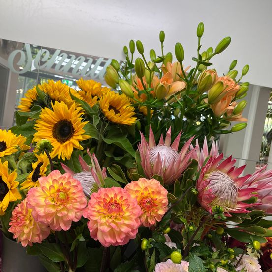 Ein lebendiger Blumenstrauß mit Sonnenblumen, Dahlien und Proteen in verschiedenen Orange- und Rosatönen, präsentiert in einer modernen Umgebung.