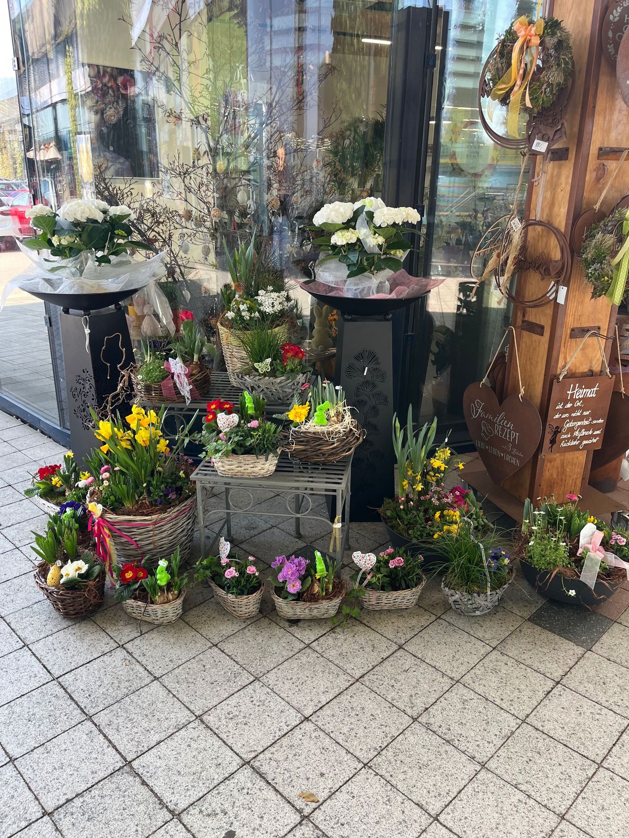 Vor einem Geschäft sind verschiedene Blumenarrangements in Weidenkörben ausgestellt, mit weißen, gelben und lila Blumen. Ein hölzerner Pfosten mit hängenden Schildern und Herzen befindet sich in der Nähe.