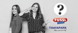 Zwei Frauen posieren für ein Foto. Eine lächelt, die andere macht ein Friedenszeichen. Im Hintergrund befindet sich ein Fragezeichen und ein Logo für FUSSL und THAYAPARK.