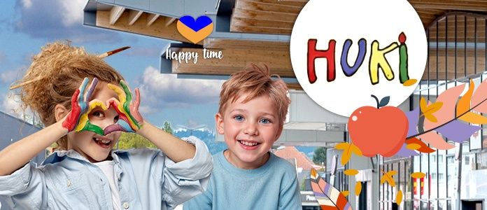 Zwei lächelnde Kinder mit Farbe an den Händen stehen in der Nähe eines Gebäudes mit dem Wort Huki. Der Himmel ist bewölkt und ein Herz und ein Apfel sind im Bild dargestellt.