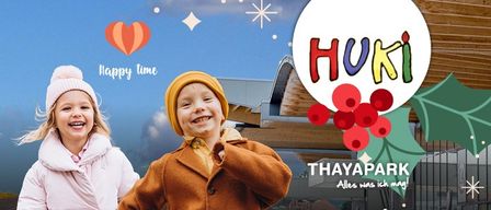 Zwei Kinder, ein Mädchen in Pink und ein Junge in Braun, lächeln vor einem Gebäude mit dem Wort 'HUKI' und einem Weihnachtsthema.