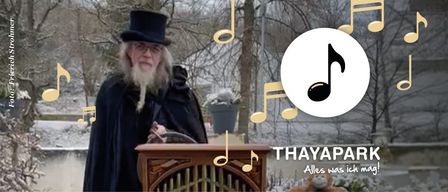 Ein alter Mann in einem Umhang und Hut steht vor einer Orgel, umgeben von Musiknoten, in einer verschneiten Landschaft. Der Text lautet 'THAYAPARK Alles was ich mag!'.