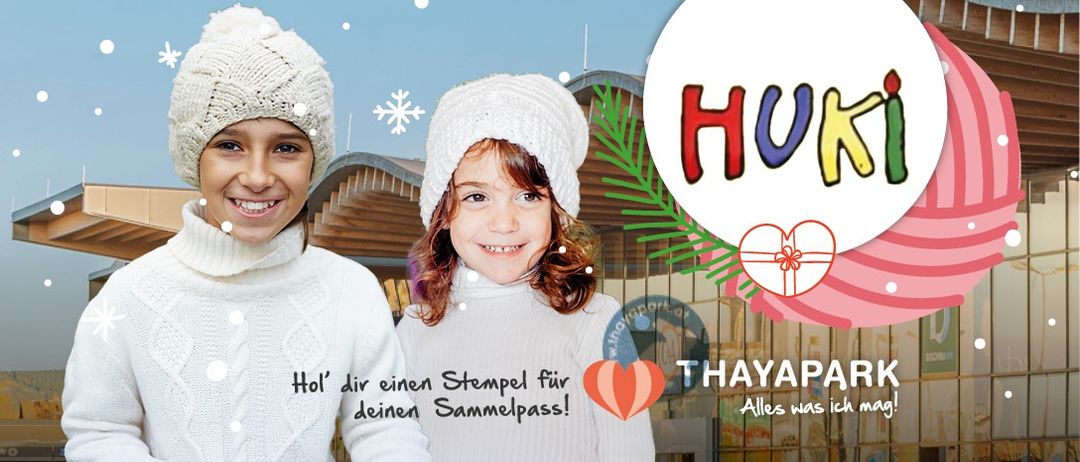 Zwei Mädchen lächeln vor einem Gebäude mit Weihnachtsdekoration, eines trägt eine Mütze und das andere eine Haube. Es gibt ein Banner mit 'HUKI', 'THAYAPARK' und einem Herzsymbol.