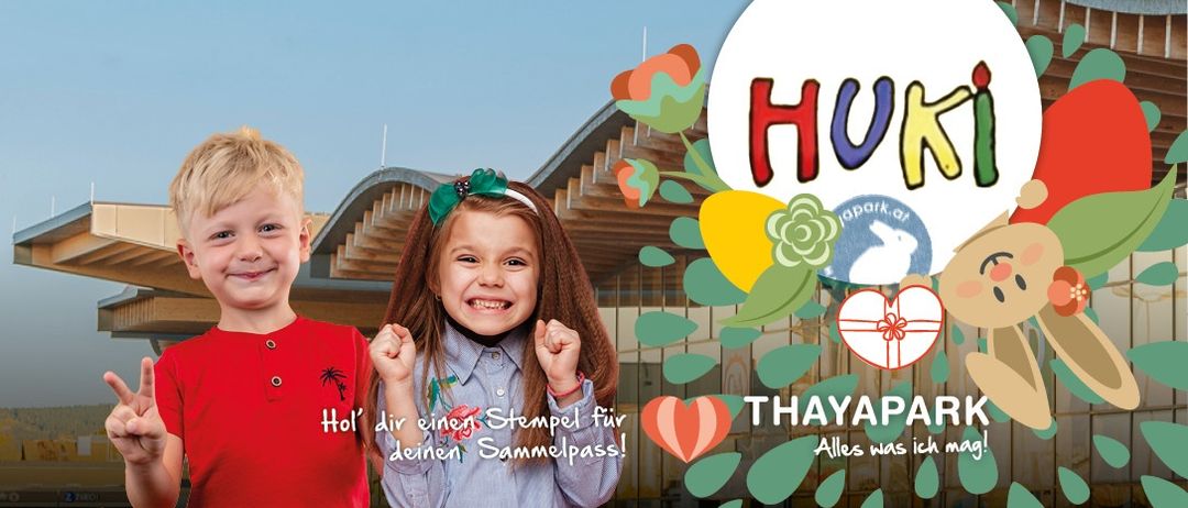 Zwei Kinder lächeln vor einem Gebäude mit Holzfassade. Der Junge trägt ein rotes Shirt, das Mädchen ein blaues Shirt mit weißer Stirnband.