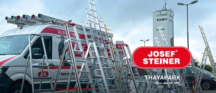 Ein weißer Van mit roten Akzenten zeigt 'Josef Steiner'. Mehrere Leitern sind dahinter gestapelt. Ein hohes weißes Gebäude und geparkte Autos sind im Hintergrund.