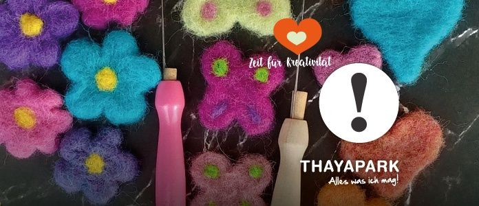 Eine Sammlung bunter Filzblumen und Herzen auf einer schwarzen Oberfläche. Zwei rosa Nadelwerkzeuge mit einem Ausrufezeichen. Der Text sagt 'Zeit für Kreativität' und 'Thayapark'.