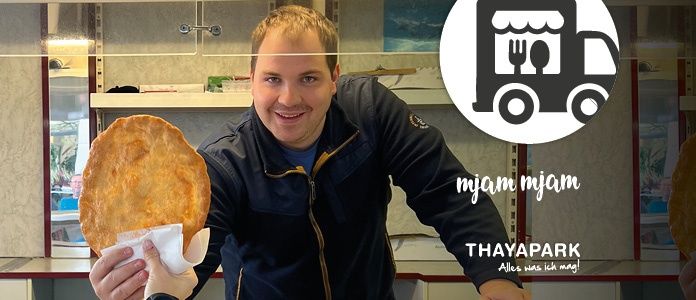 Ein Mann in einem schwarzen Jackenmantel hält ein großes rundes Essensstück in einer Küche. Der Mann lächelt. Im Hintergrund befindet sich ein weißes Logo mit den Worten 'myam myam'.