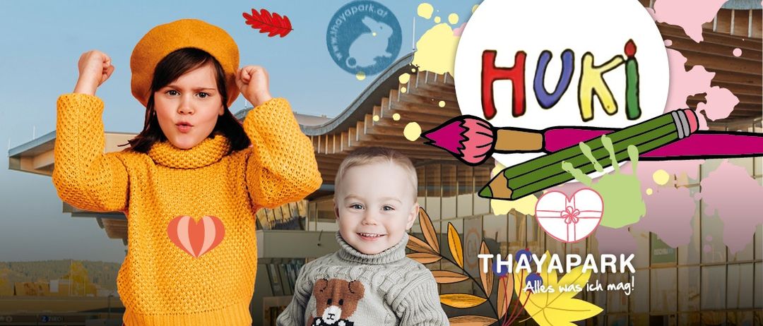 Zwei Kinder, ein Mädchen in einem gelben Pullover und ein Junge in einem grauen Pullover mit einem Bärendesign, lächeln vor einem Gebäude mit Herbstdekorationen. Das Wort 'Huki' ist in bunten Buchstaben prominent angezeigt.