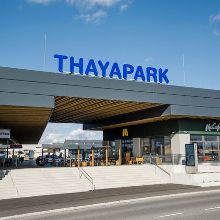 Außenansicht des Thaypark Einkaufszentrums unter einem blauen Himmel. Der Eingang zeigt ein blaues Schild mit dem Wort Thaypark, und ein McDonald's Restaurant ist auf der rechten Seite zu sehen.