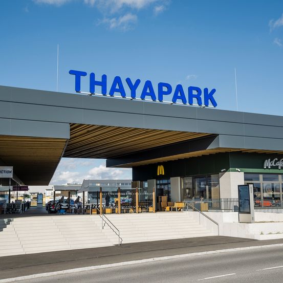 Außenansicht des Thaypark Einkaufszentrums unter einem blauen Himmel. Der Eingang zeigt ein blaues Schild mit dem Wort Thaypark, und ein McDonald's Restaurant ist auf der rechten Seite zu sehen.