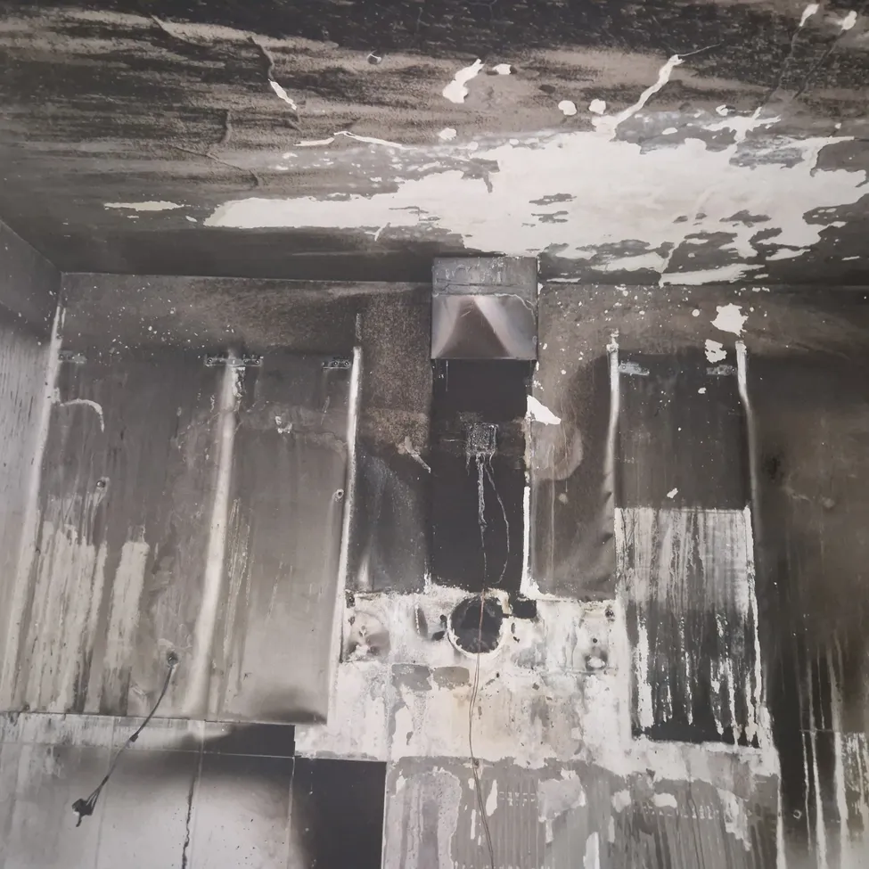 Das Bild zeigt einen Raum mit Feuerschäden an Wänden und Decke. Ruß- und Rauchspuren bedecken die Oberflächen, und es befindet sich ein metallischer Lüftungsschacht an der Decke. Kabel hängen von der Decke, und in der Wand ist ein Loch sichtbar.