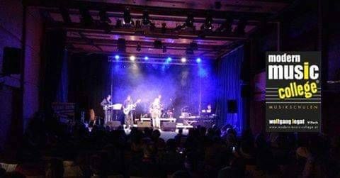 A live performance on stage with musicians playing guitars and keyboards, under blue and purple lights. The audience is seated in front of the stage, watching the show. The stage has a banner with the text 'modern music college'.