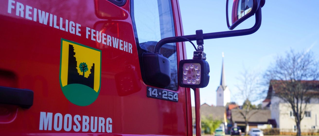 Nahaufnahme der Seite eines roten Feuerwehrwagens aus Moosburg mit Logo und Scheinwerfer. Im Hintergrund sind eine Kirche, Autos und ein klarer Himmel zu sehen.