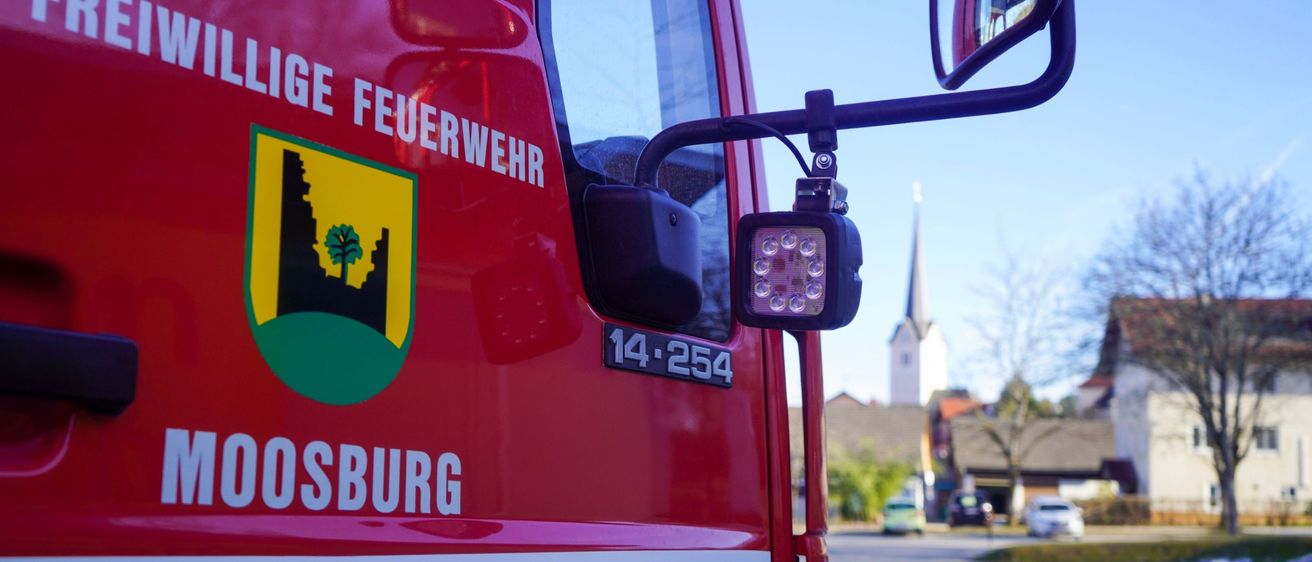 Nahaufnahme der Seite eines roten Feuerwehrwagens aus Moosburg mit Logo und Scheinwerfer. Im Hintergrund sind eine Kirche, Autos und ein klarer Himmel zu sehen.