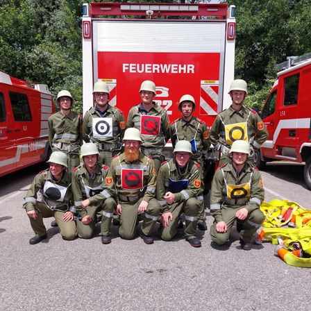 Eine Gruppe Feuerwehrleute posiert mit Helmen für ein Foto vor einem Feuerwehrauto. Sie tragen alle Uniformen, und einige haben Ziele auf der Brust.
