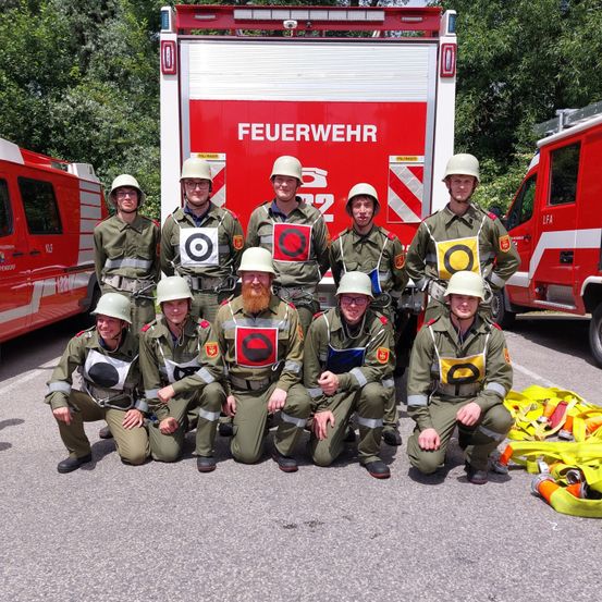 Eine Gruppe Feuerwehrleute posiert mit Helmen für ein Foto vor einem Feuerwehrauto. Sie tragen alle Uniformen, und einige haben Ziele auf der Brust.